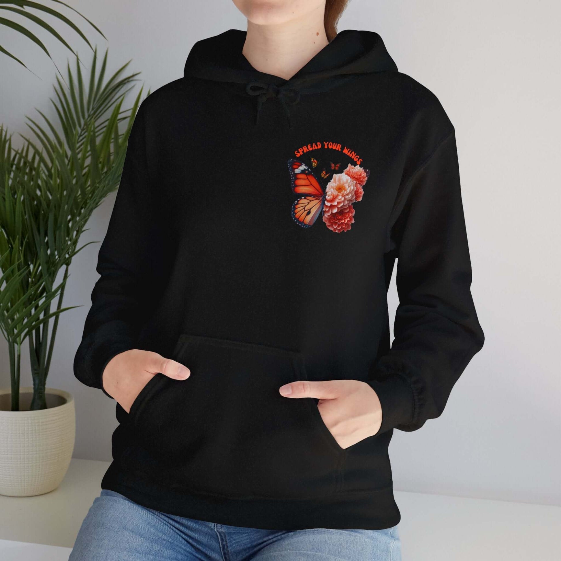 Person wearing "Spread Your Wings" butterfly and flowers hoodie in black, showcasing vibrant butterfly and floral design with hands in pockets.