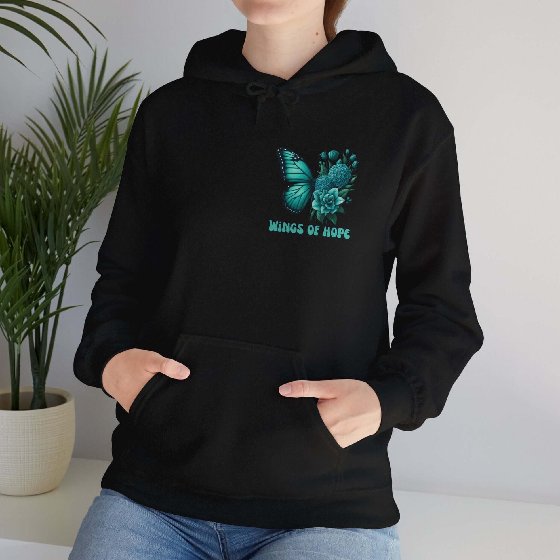 Woman wearing "Wings Of Hope" Butterfly and Flowers Hoodie, standing by a potted plant.