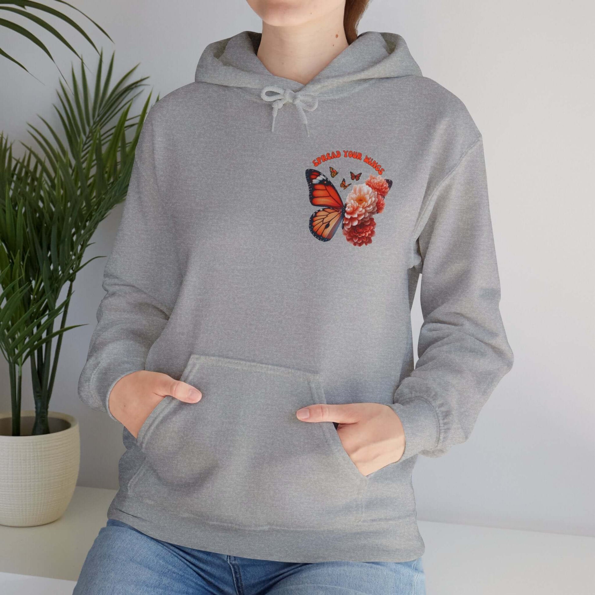 Woman wearing "Spread Your Wings" butterfly and flowers hoodie with butterfly and floral design, standing indoors near a plant