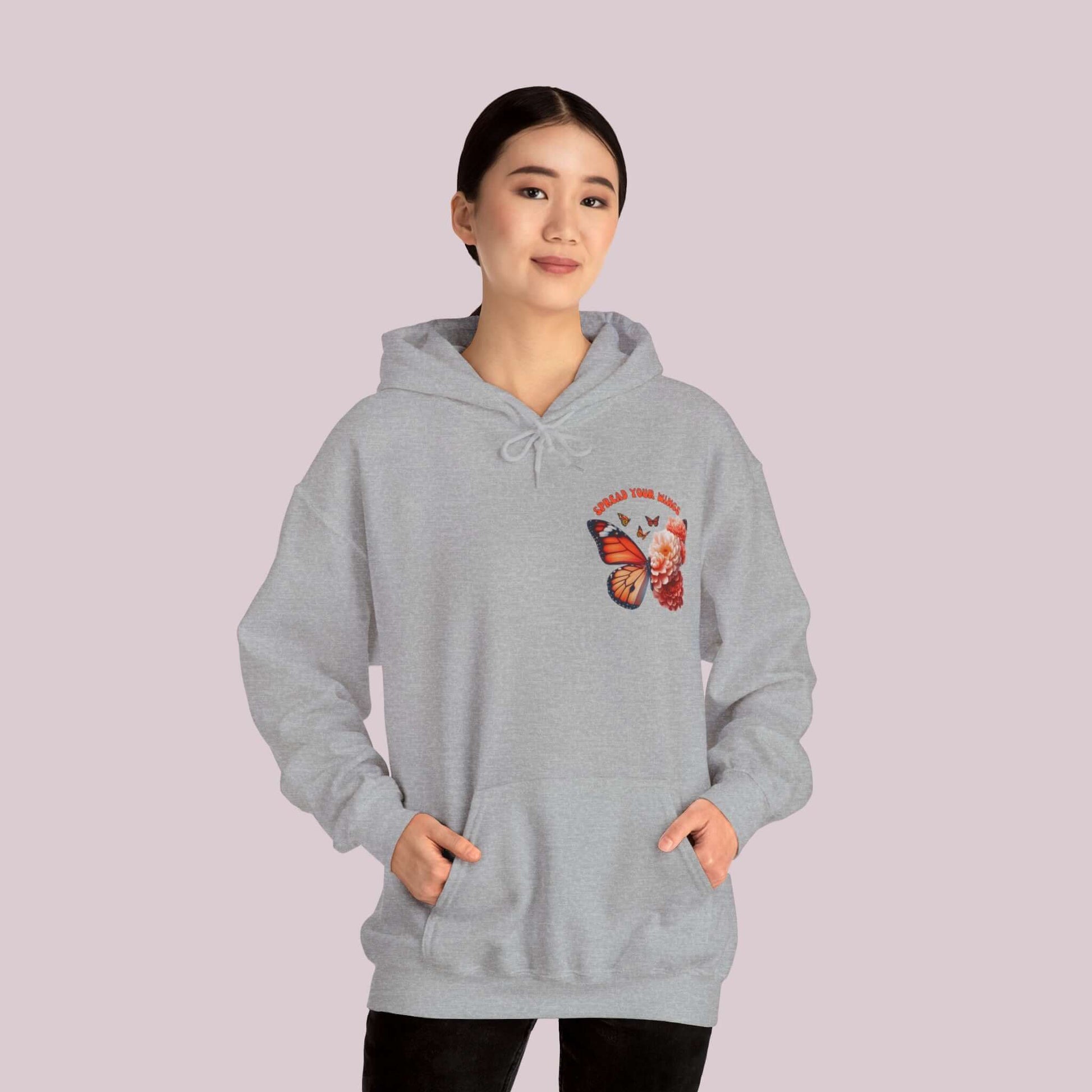 Woman wearing gray "Spread Your Wings" butterfly and flowers hoodie with hands in pockets.